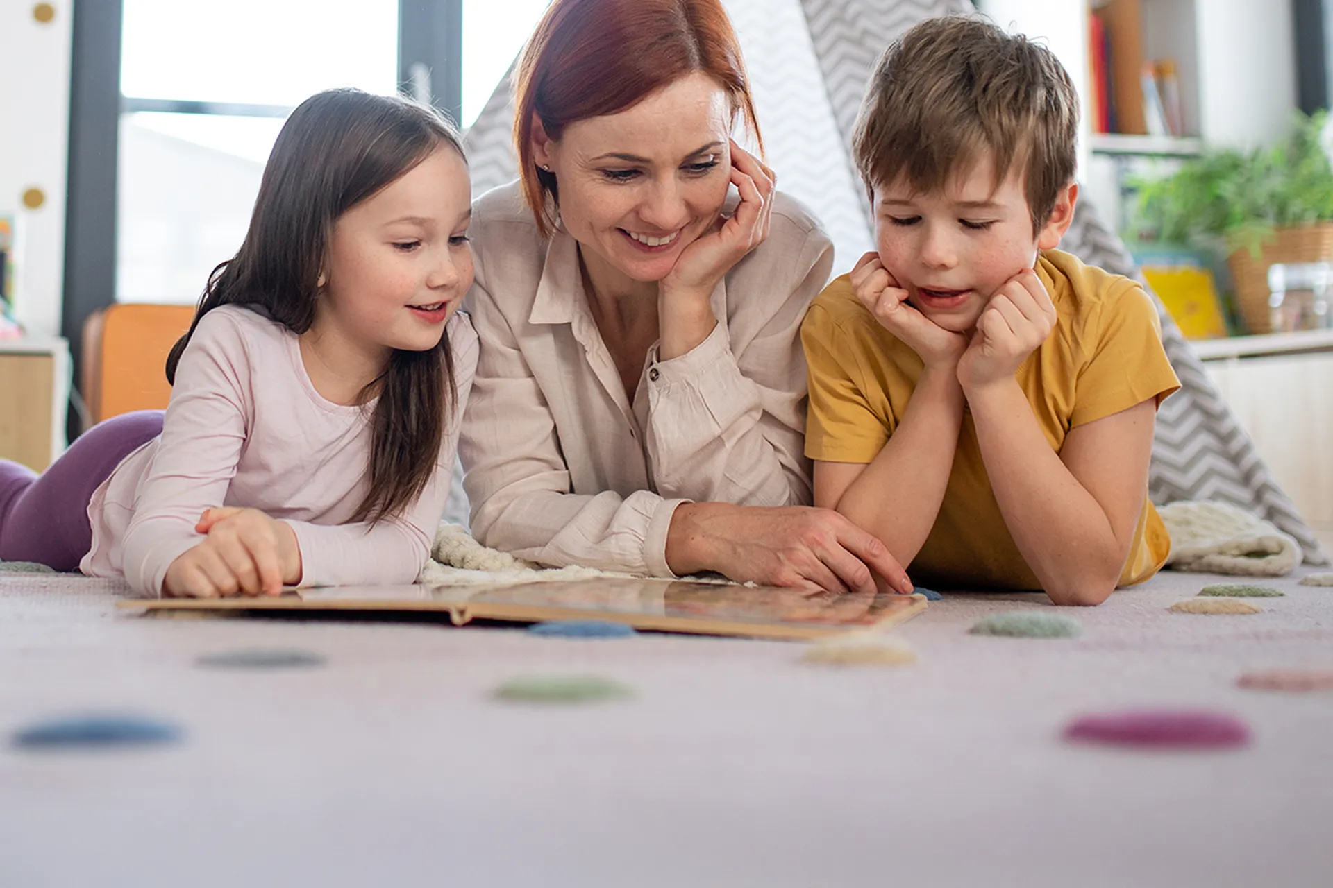 Eine Mutter liegt mit ihren zwei Kindern auf dem Boden und liest ihnen aus einem Buch vor