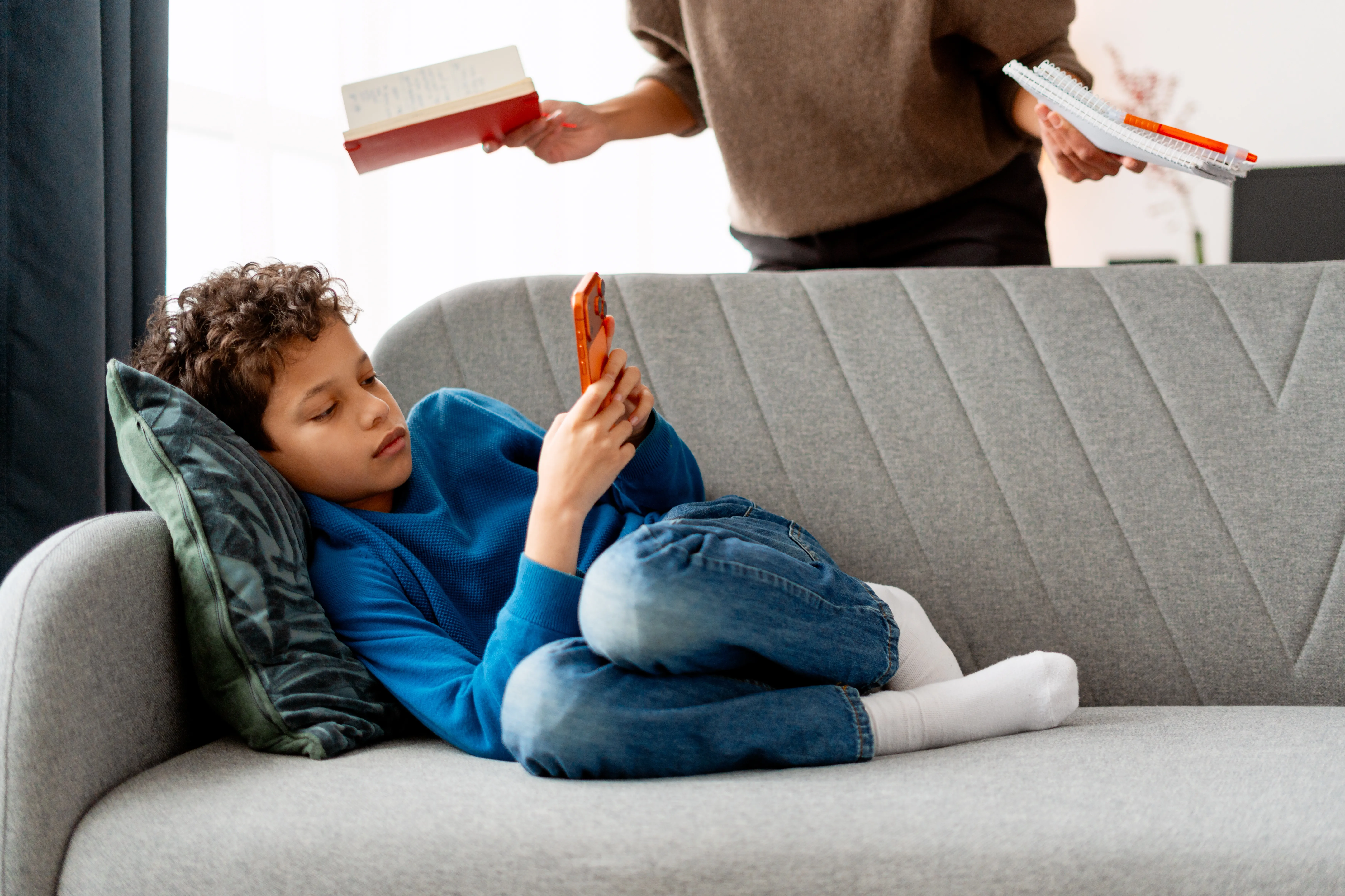 Kind liegt mit Handy auf der Couch und ignoriert Mutter mit Buechern und SChulaufgaben