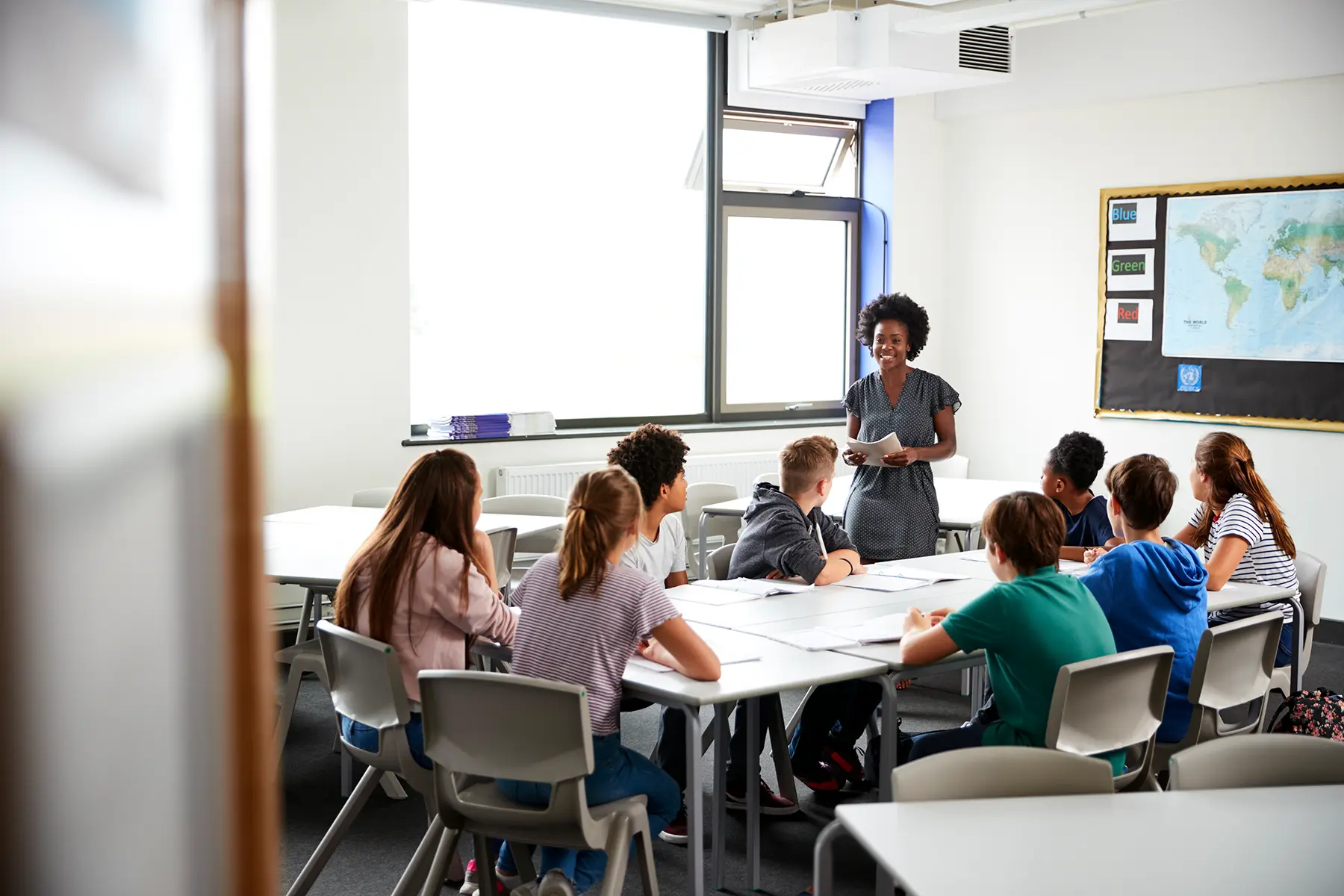 Kinder sitzen in einem Klassenzimmer an einem Tisch, Lehrerin erklaert etwas; Linkziel: Kategorie Schule