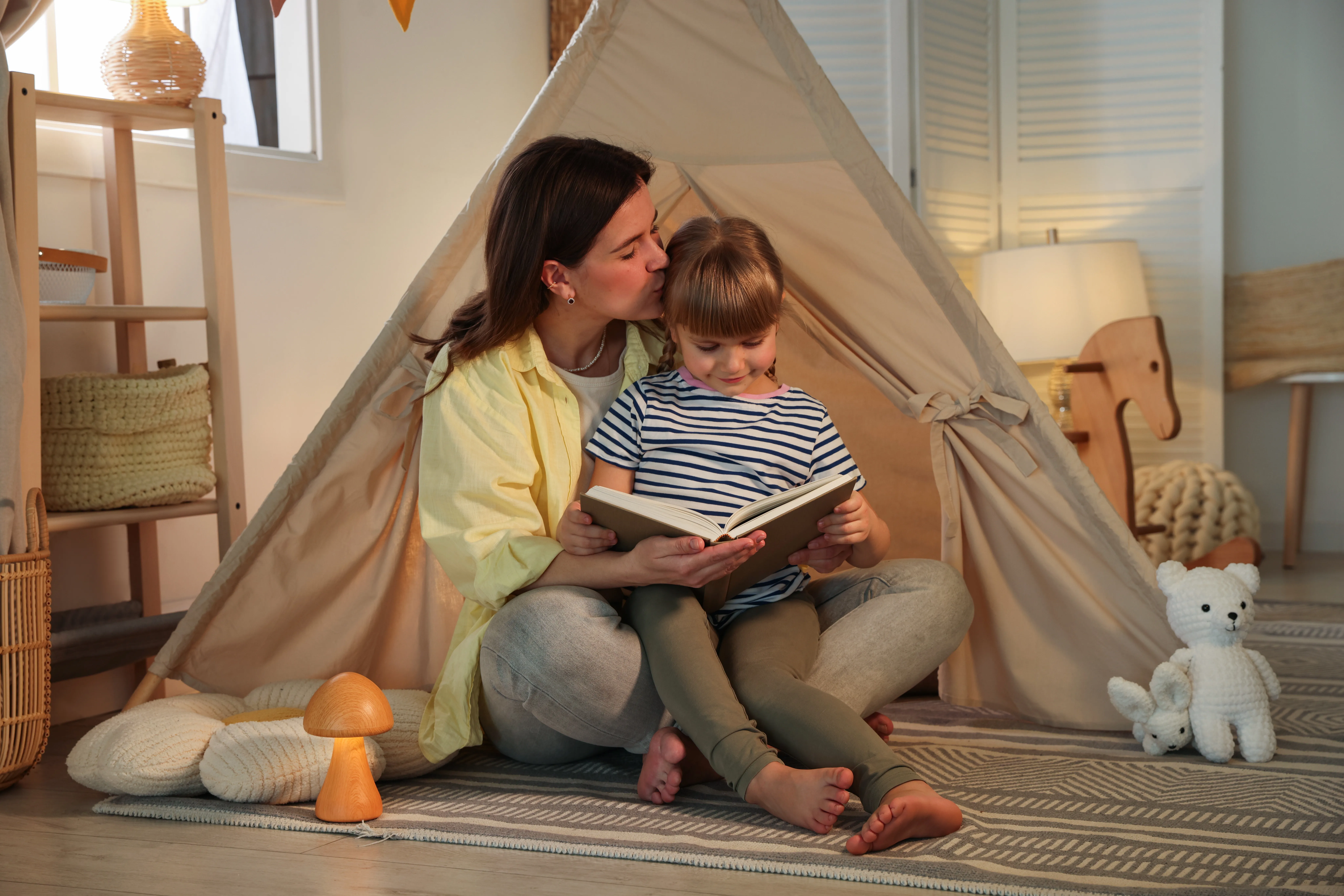 Mutter sitzt mit Kind vor einem Zelt, das im Zimmer aufgebaut wurde und liest ein Buch vor;